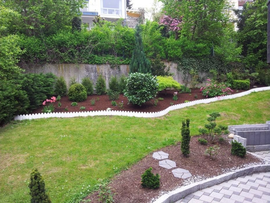 Ein gepflegter Hinterhofgarten mit Rasen, mit Mulch bedeckten Blumenbeeten, einem weißen Begrenzungszaun und einer mit Steinen gepflasterten Terrasse.