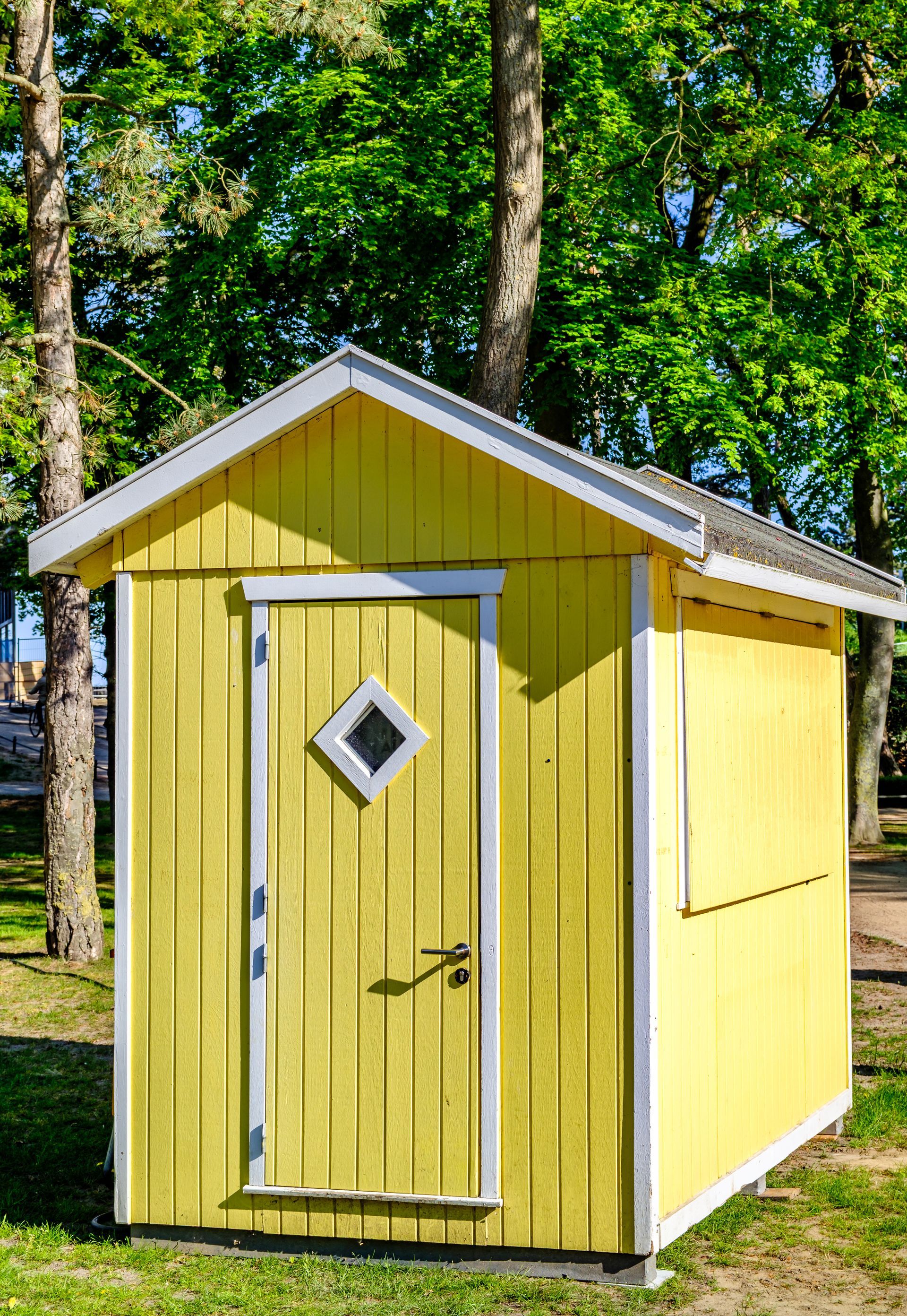 Abri jaune avec des bordures blanches, une fenêtre en forme de losange et un arbre vert en arrière-plan.