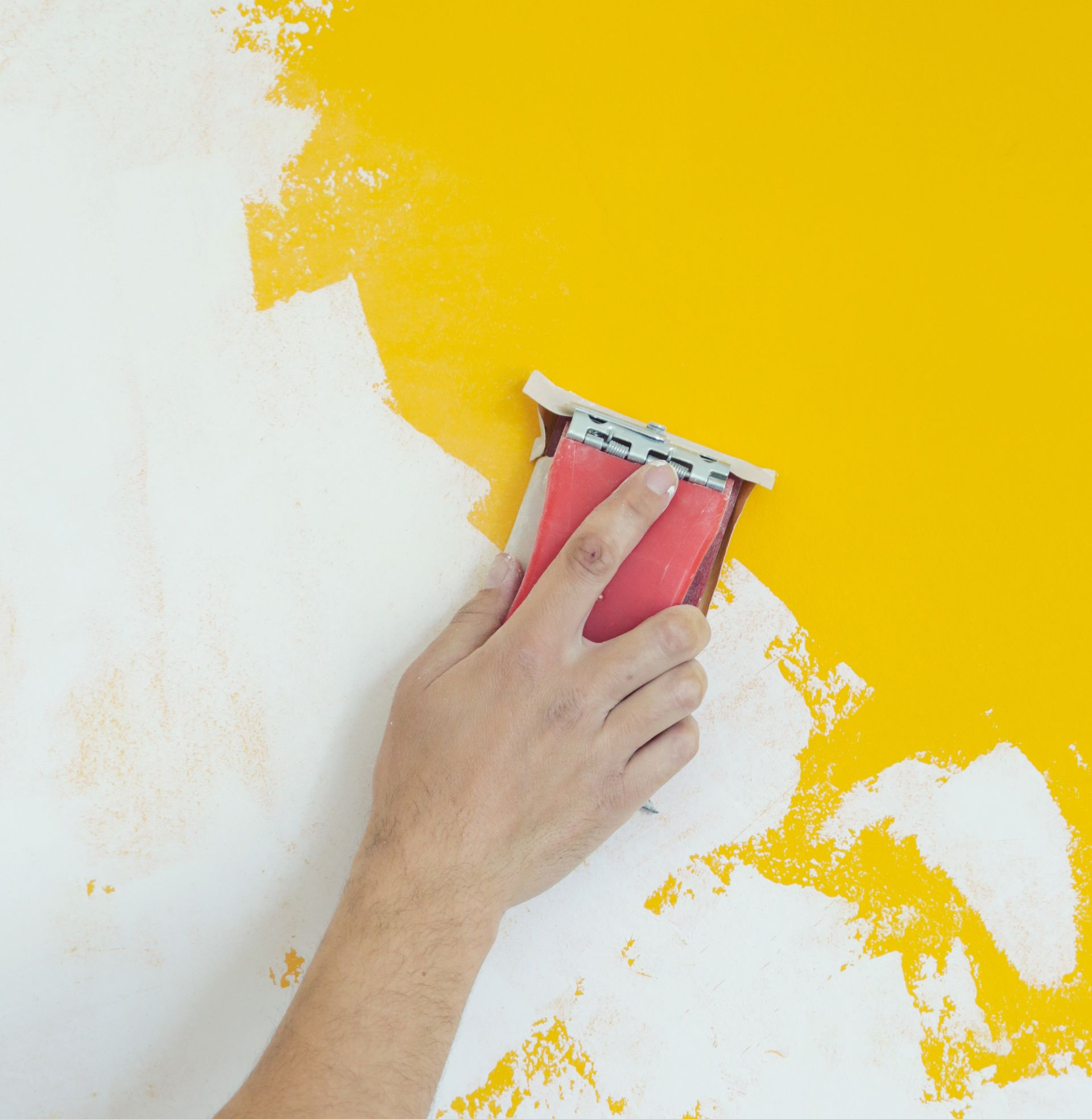 Main utilisant un bloc de ponçage sur un mur peint en blanc et jaune.