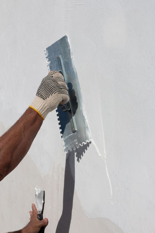 Une personne utilise une truelle crantée pour appliquer un revêtement blanc sur un mur, portant un gant de travail.