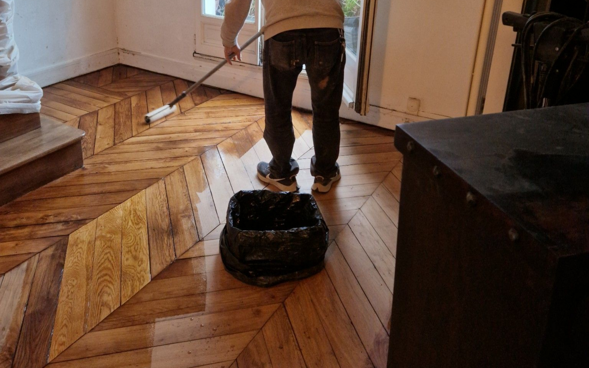Une personne applique un produit d'étanchéité sur un parquet à l'aide d'un rouleau. Un sac-poubelle et une armoire en bois foncé se trouvent à proximité.