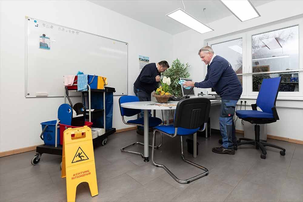 Zwei Männer reinigen einen Konferenzraum. Auf dem Boden liegt ein gelbes Warnschild.