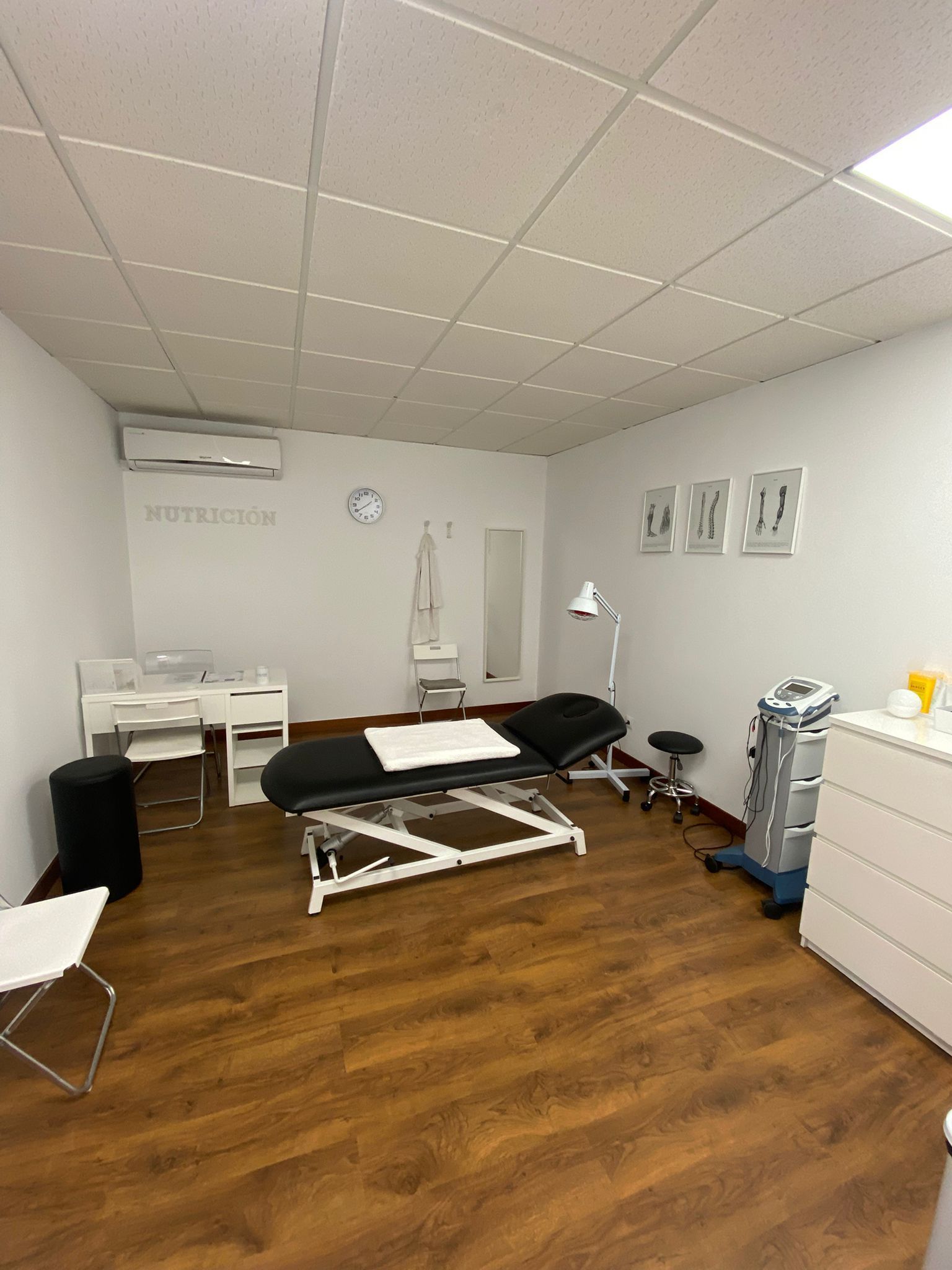 Una sala de tratamiento limpia y moderna con camilla de masaje, muebles blancos, equipo médico y suelo de madera clara.