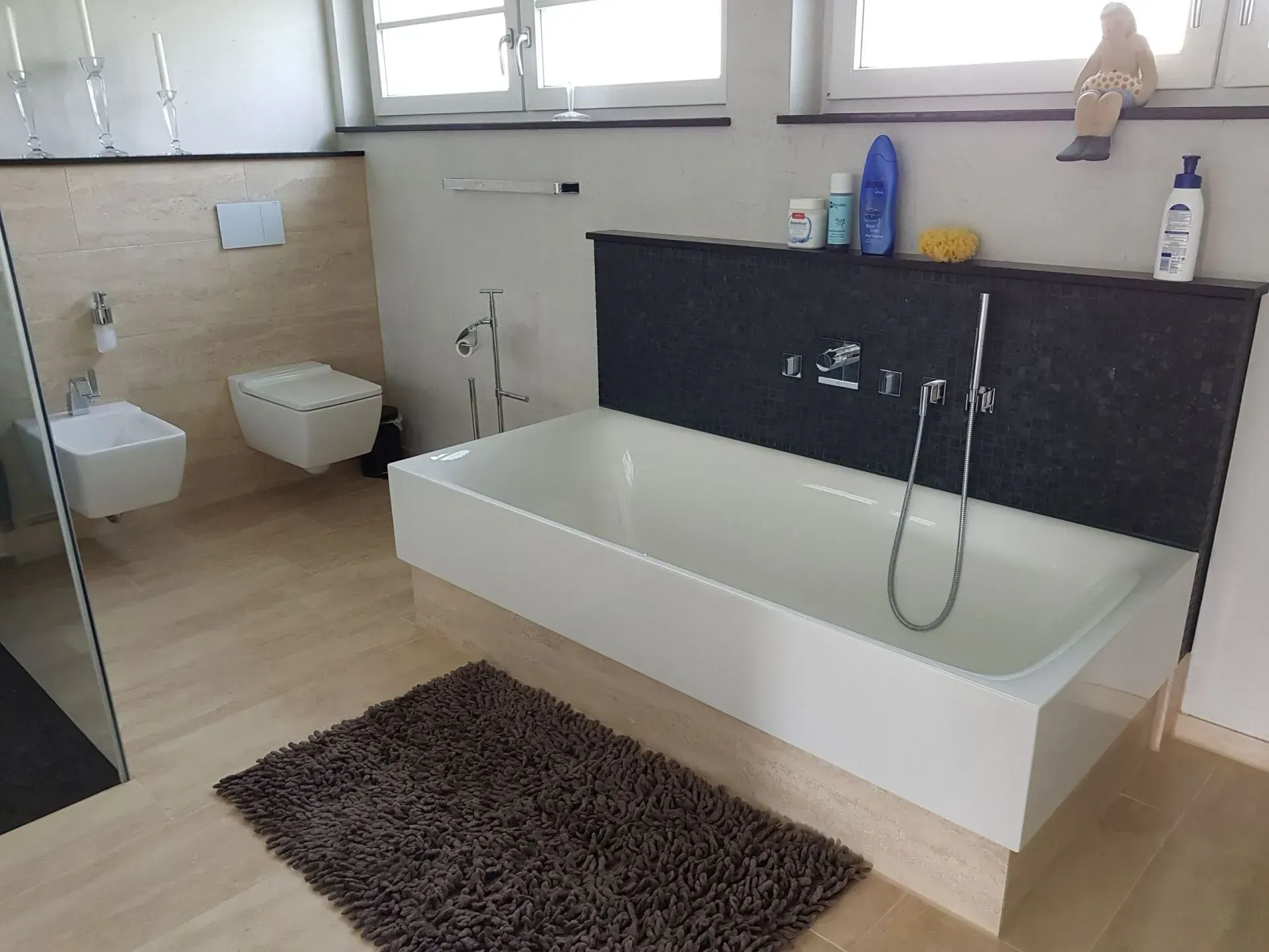 Modernes Badezimmer mit weißer Badewanne, Toilette, Waschbecken und brauner Badematte unter einem Fenster.