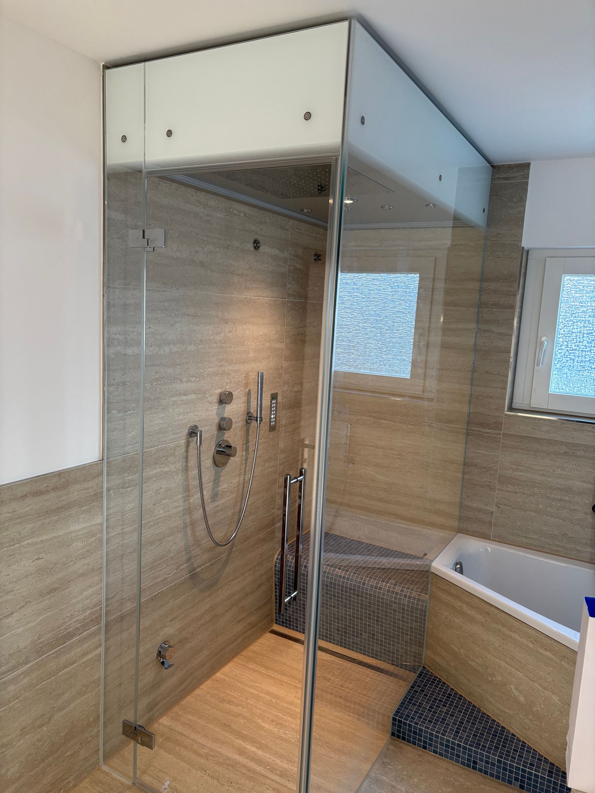 Modernes Badezimmer mit Glasdusche, beigefarbenen Fliesen und einer weißen Badewanne unter kleinen Fenstern.