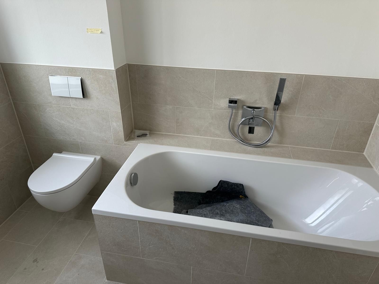 Modernes Badezimmer mit weißer Badewanne, wandhängendem WC und beigefarbenen Fliesenwänden; Kleidung in der Badewanne