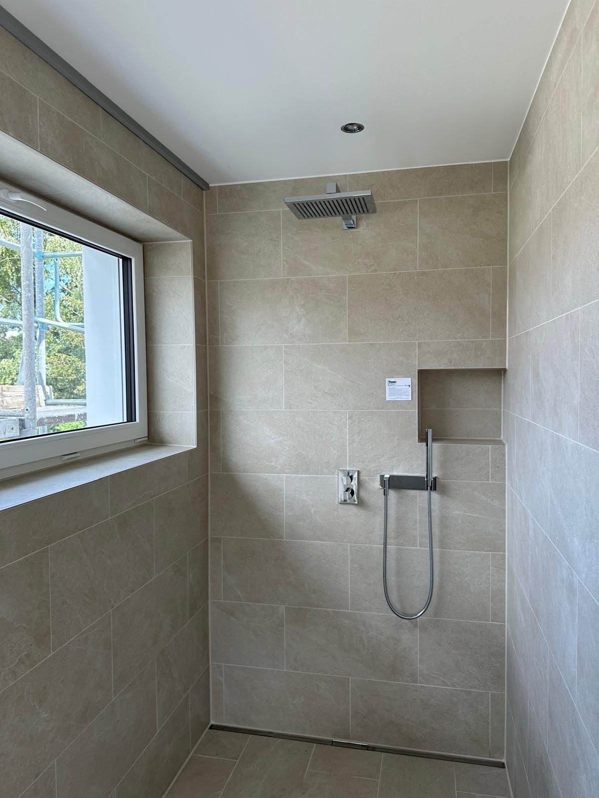 Leere, geflieste Dusche mit wandmontiertem Duschkopf und Handbrause neben einem Fenster