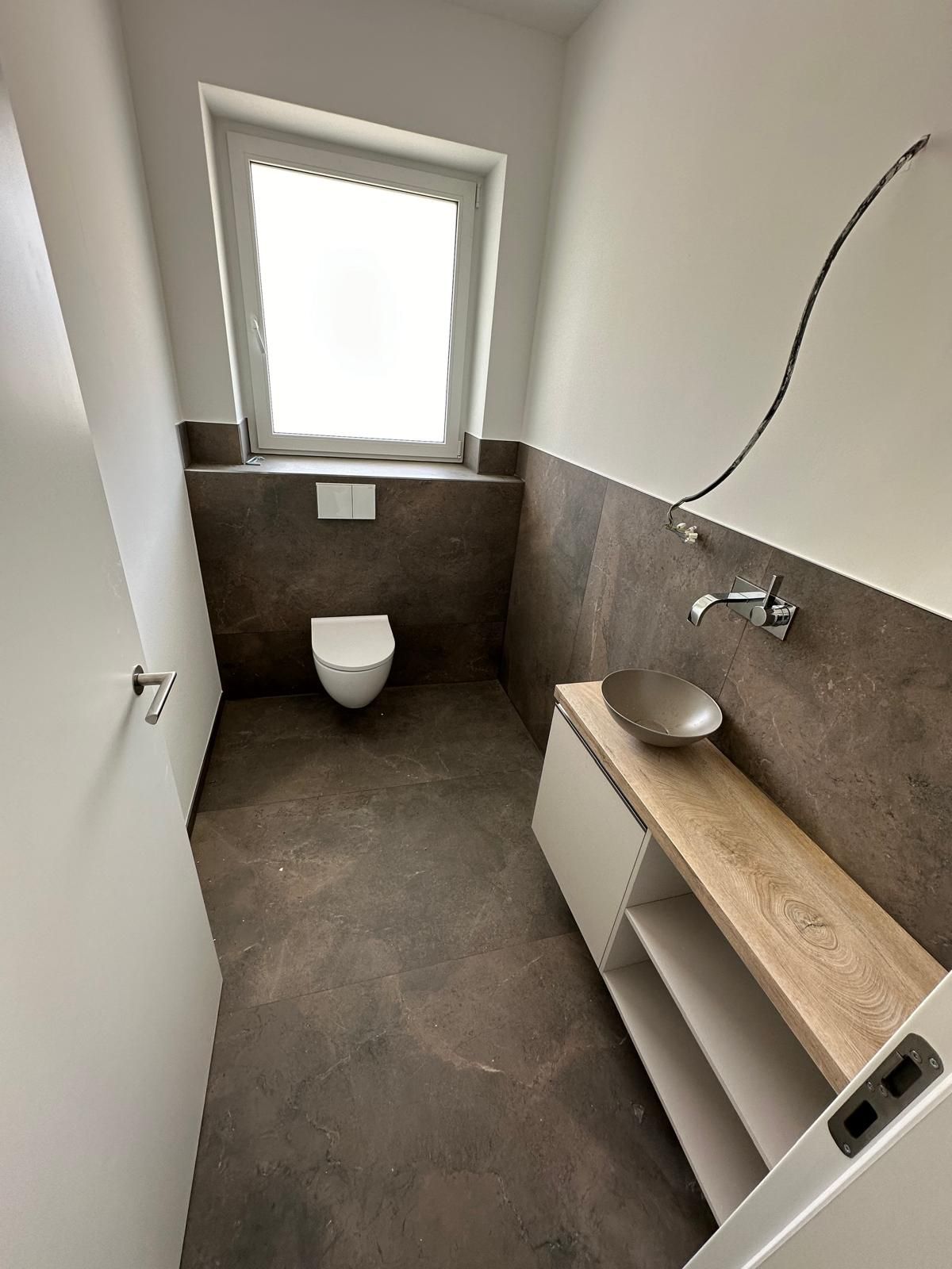 Schmale, moderne Badezimmerausstattung mit Toilette, Waschbecken, Fenster und braun gefliesten Wänden und Boden.