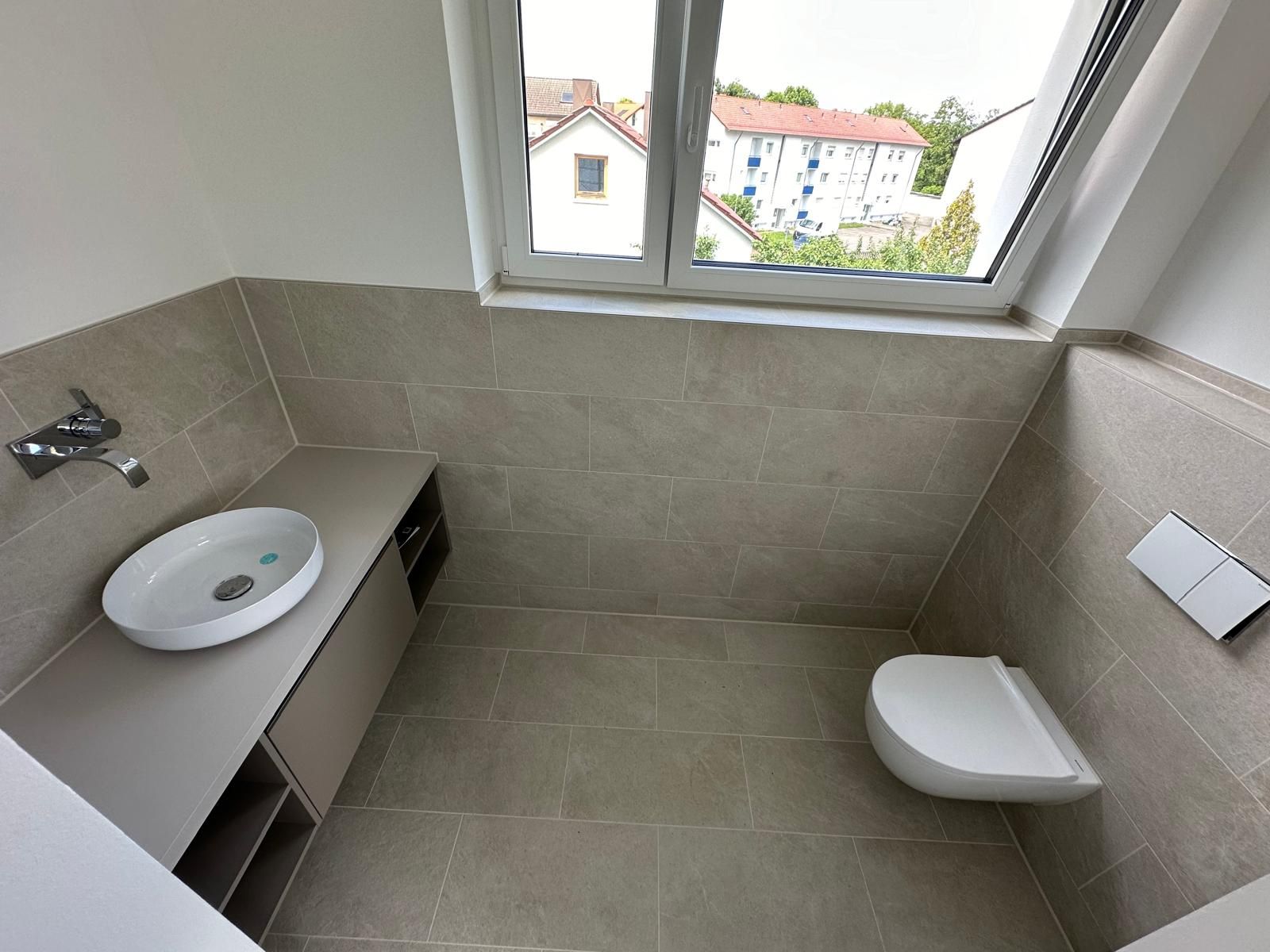 Kleines gefliestes Badezimmer mit wandmontiertem Waschbecken, Toilette und Fenster über einer schrägen Wand.