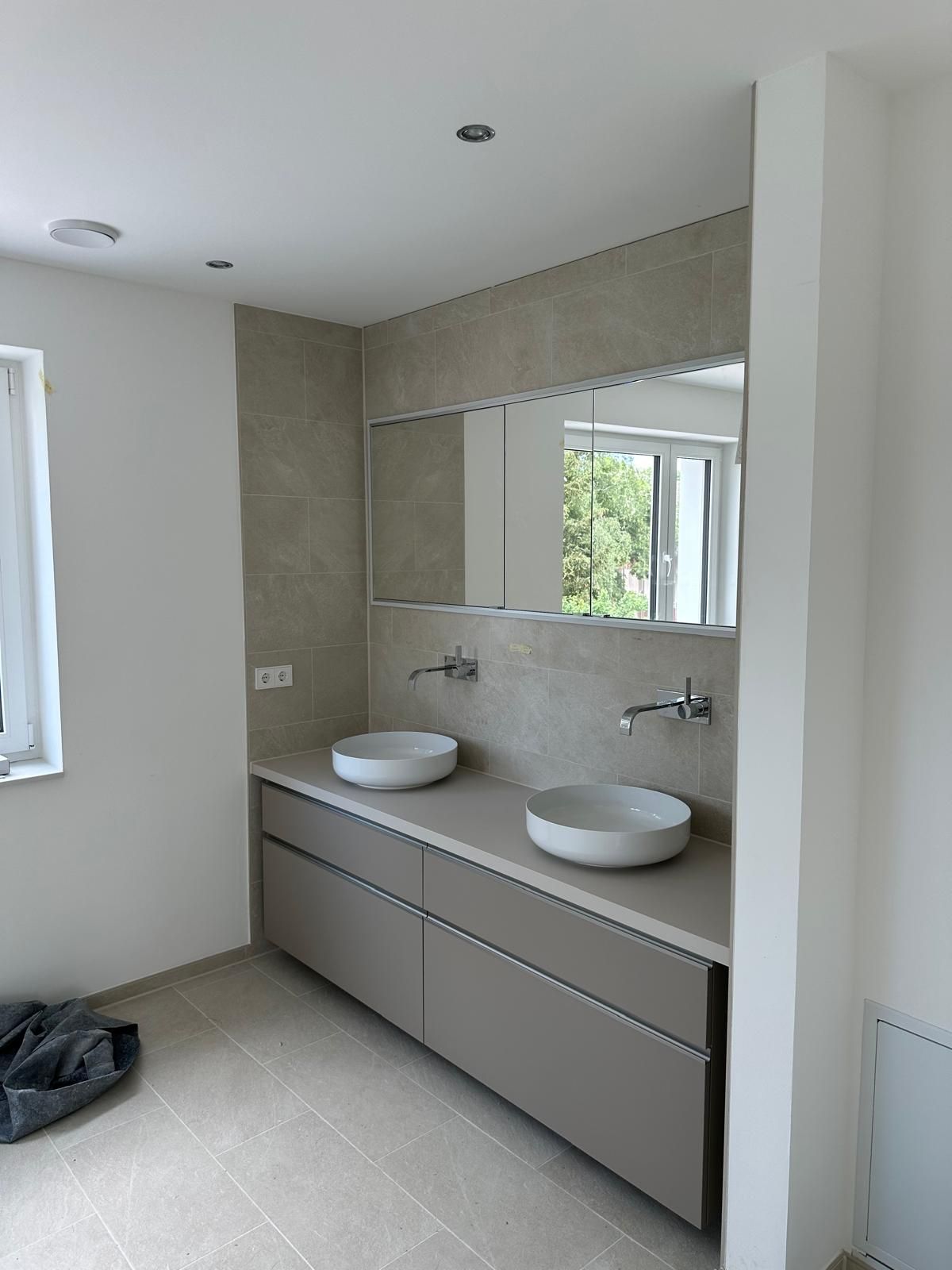 Modernes Badezimmer mit Doppelwaschbecken, großem Spiegel, grauen Schränken und einem Fenster.