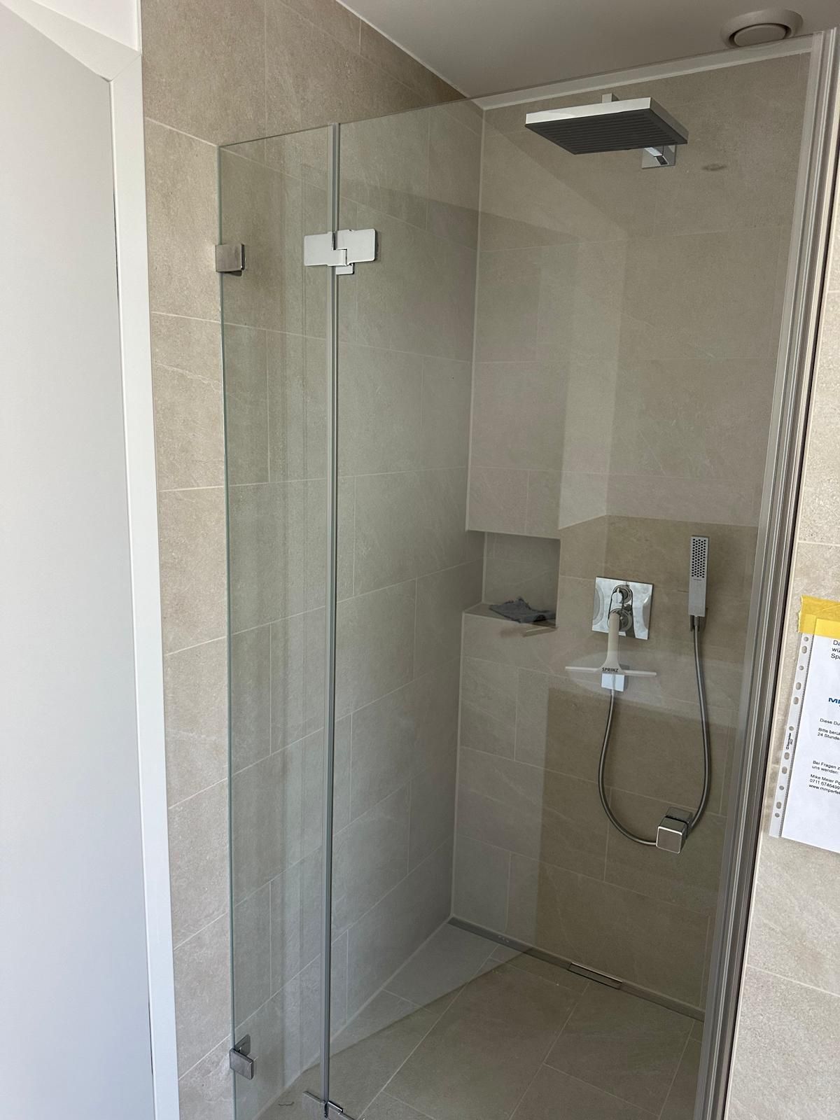 Kleine, moderne Duschkabine mit Glastür, Handbrause und beige gefliesten Wänden.