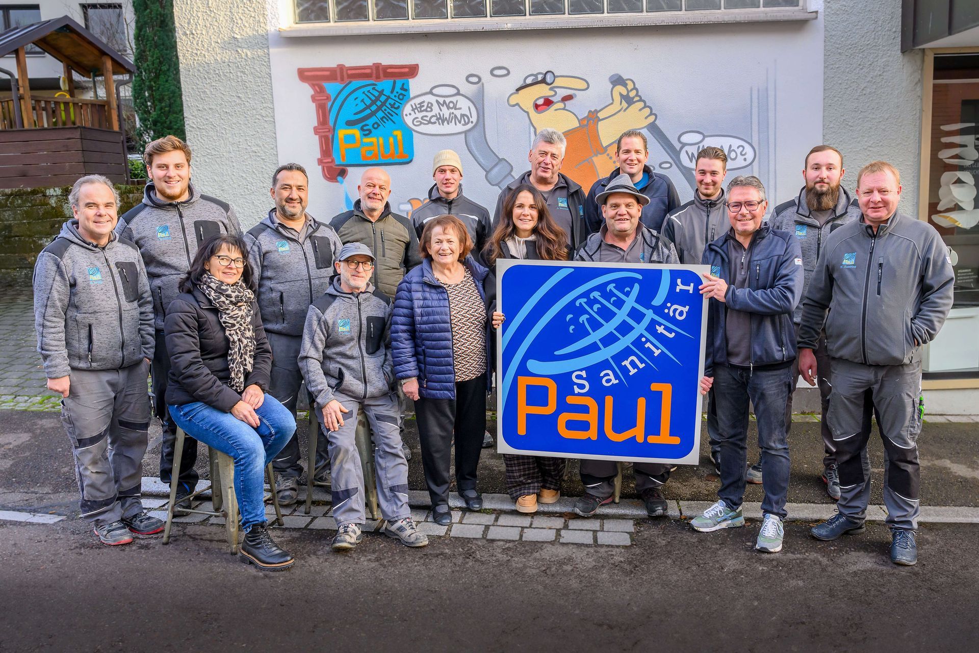 Sanitär Paul Mitarbeiter posieren vor dem Firmengebäude und halten das Firmenlogo als Schild in der Hand.