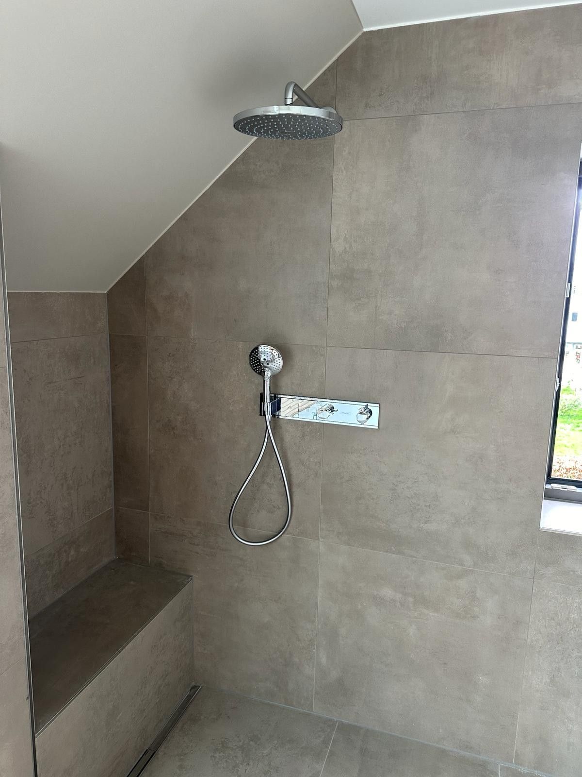Moderne, geflieste Dusche mit Regenduschkopf, Handbrause und eingebauter Sitzbank unter einer Dachschräge