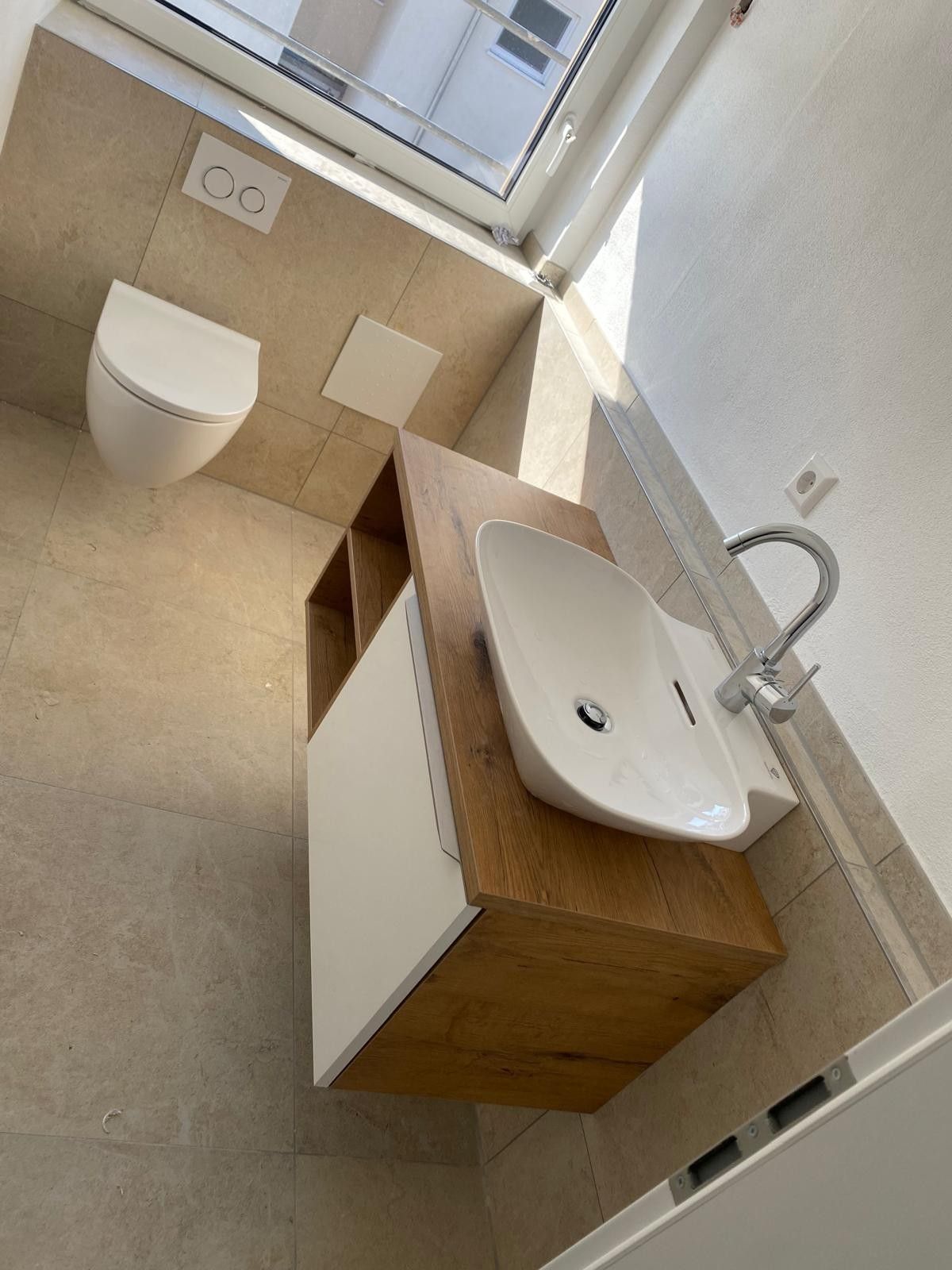 Kleines Badezimmer mit wandhängender Toilette und hölzernem Waschtisch mit weißem Waschbecken neben einem Fenster.