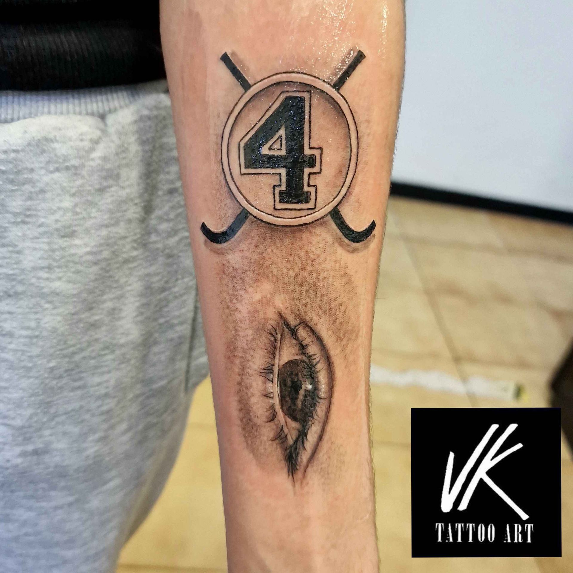 Tatuaje de un emblema con temática de hockey con el número 4, palos de hockey cruzados y un ojo en el brazo de una persona.