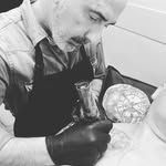 Un hombre con barba, con guantes y delantal, tatuando a una persona sobre una mesa.