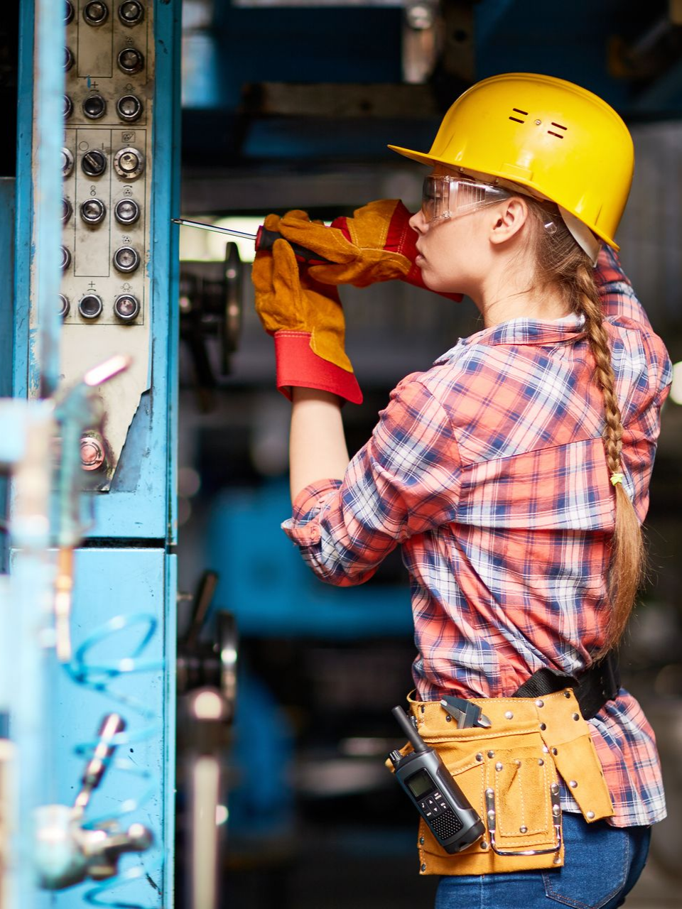 Femme en tenue de sécurité, travaillant sur des machines, portant des gants, un casque et des lunettes de protection.