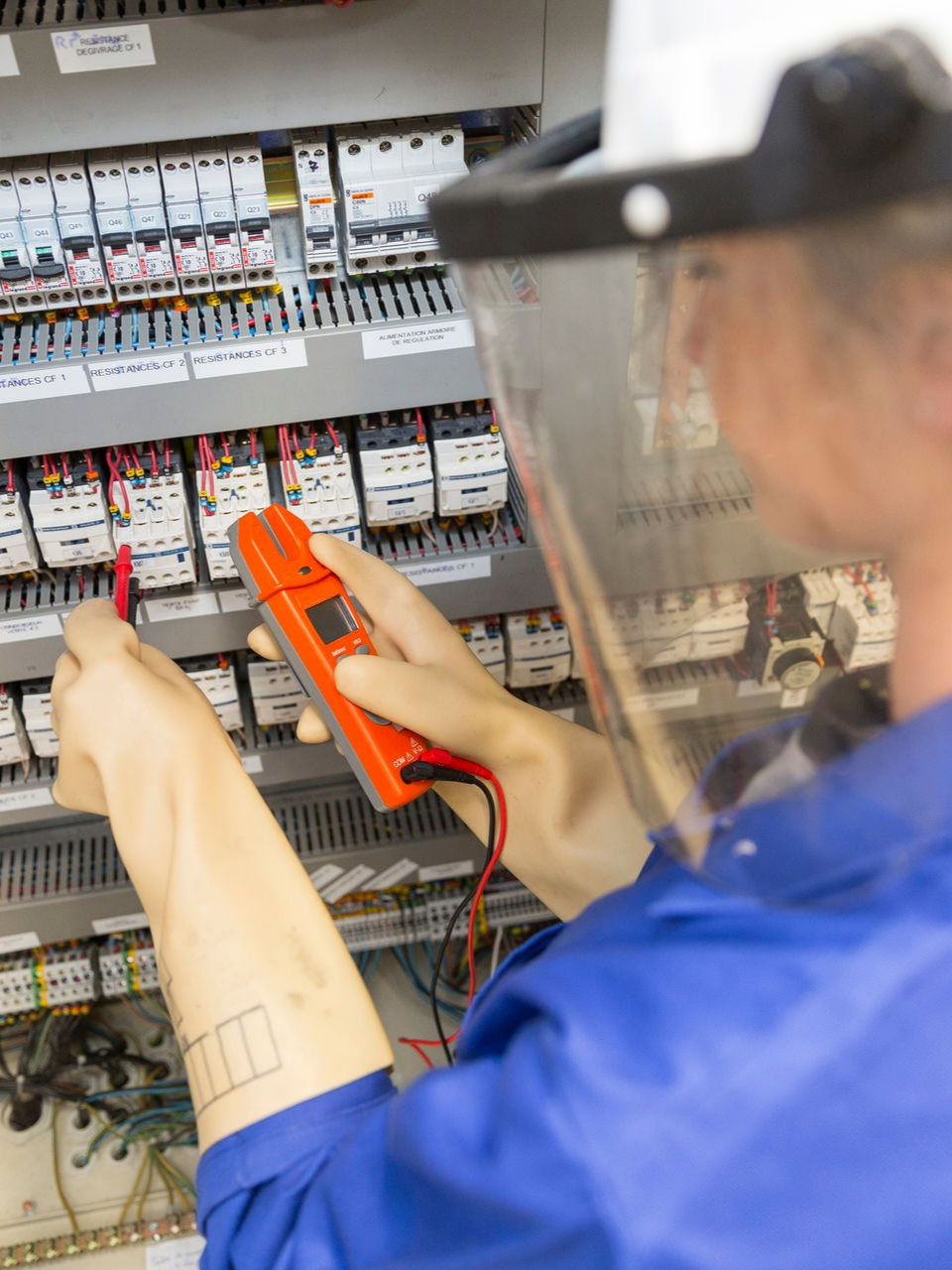 Un électricien en combinaison bleue teste un panneau de commande avec un multimètre ; il porte une visière de protection.