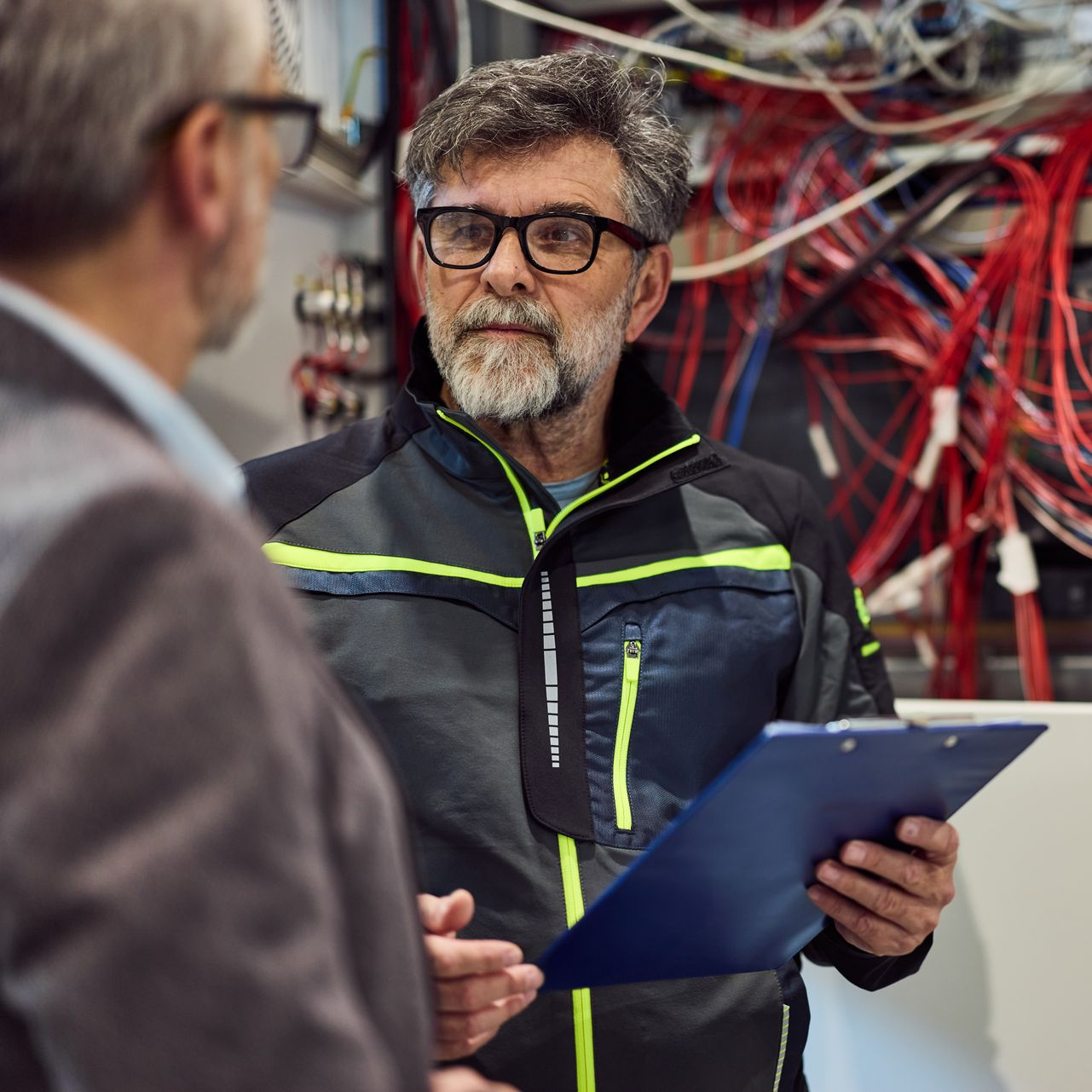 Deux hommes devant des câbles électriques, l'un d'eux tenant un bloc-notes.