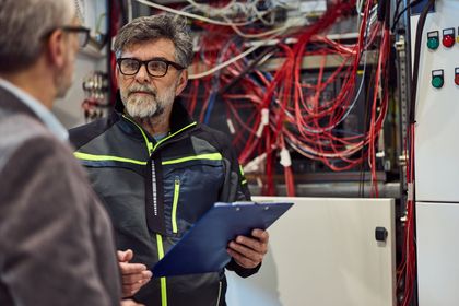 Deux hommes, dont l'un tient un bloc-notes, examinent le câblage électrique dans une salle de panneaux de commande.