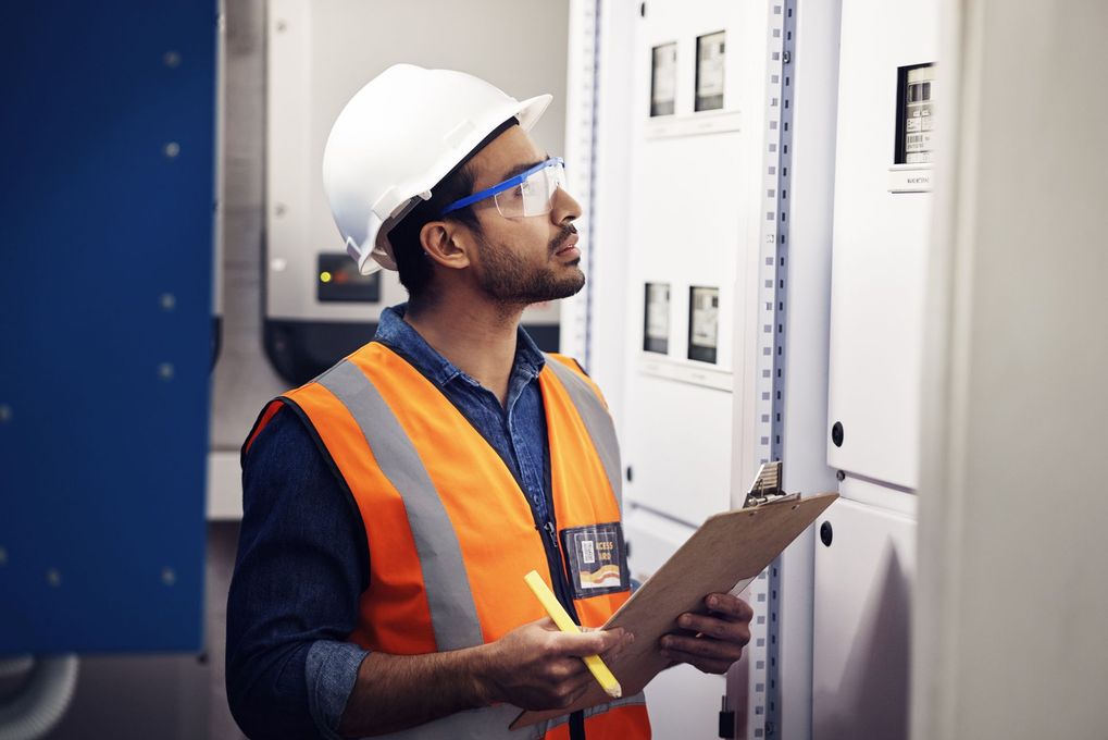 Un électricien en tenue de sécurité inspecte un tableau électrique, tenant un bloc-notes.