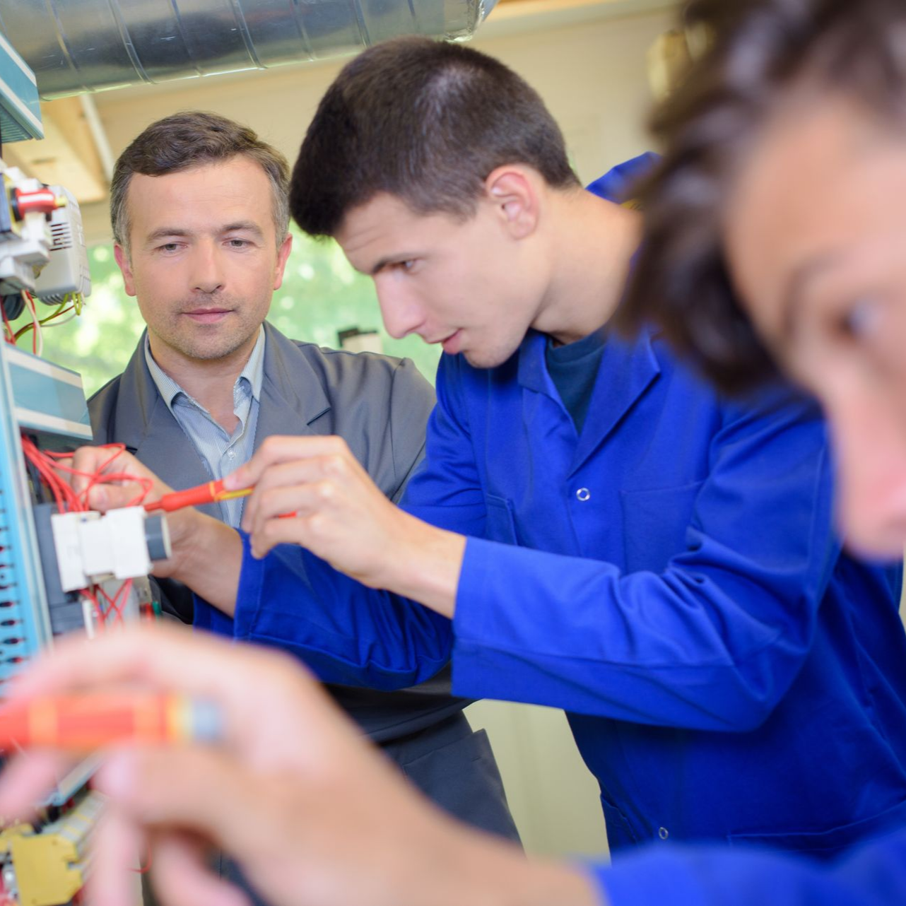 Un homme et deux autres personnes travaillent sur des câbles électriques. Les hommes portent des combinaisons bleues et l'autre un costume. À l'intérieur.