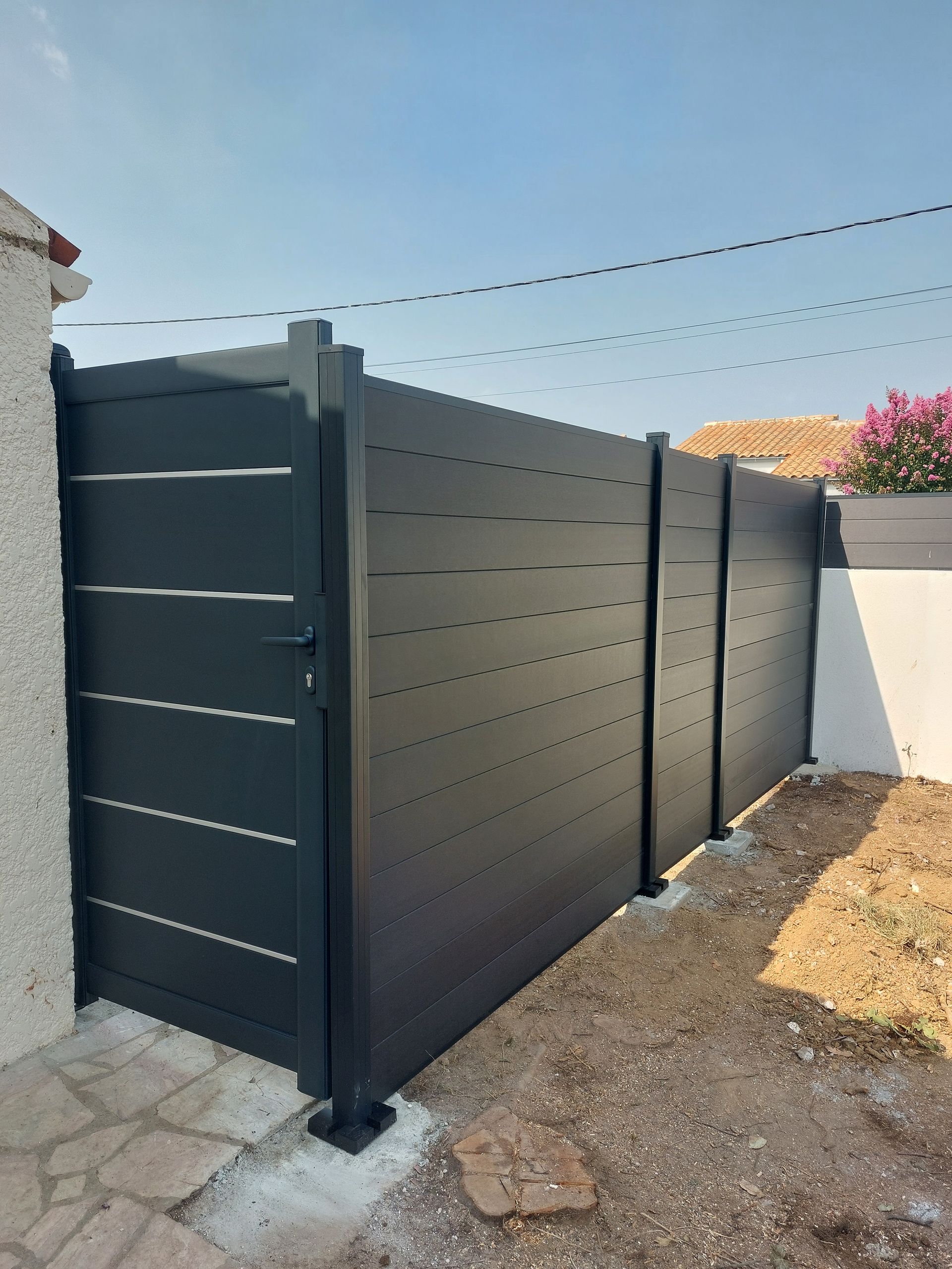 Une clôture d'intimité noire mate à lattes horizontales, installée sur une terrasse en pierre à côté d'un mur blanc.