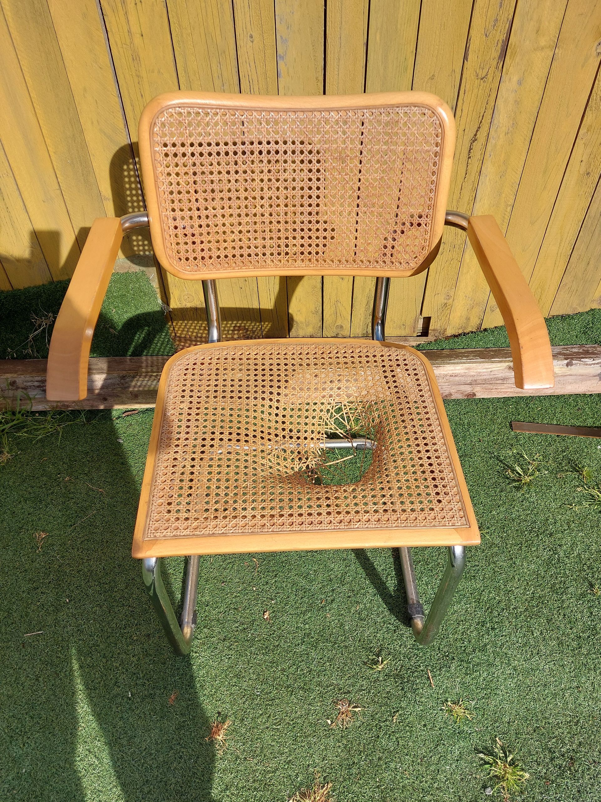 Une chaise vintage à structure chromée, avec des accoudoirs en bois clair et une assise et un dossier en rotin tressé abîmés.