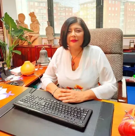 Mujer sentada en un escritorio con teclado, sonriendo en una oficina luminosa cerca de una ventana.