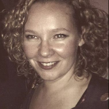 Frau mit lockigem blondem Haar, lächelnd, trägt dunkles Oberteil.