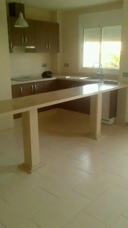 Cocina luminosa con una isla blanca, armarios marrones, suelo de baldosas y luz natural de la ventana.