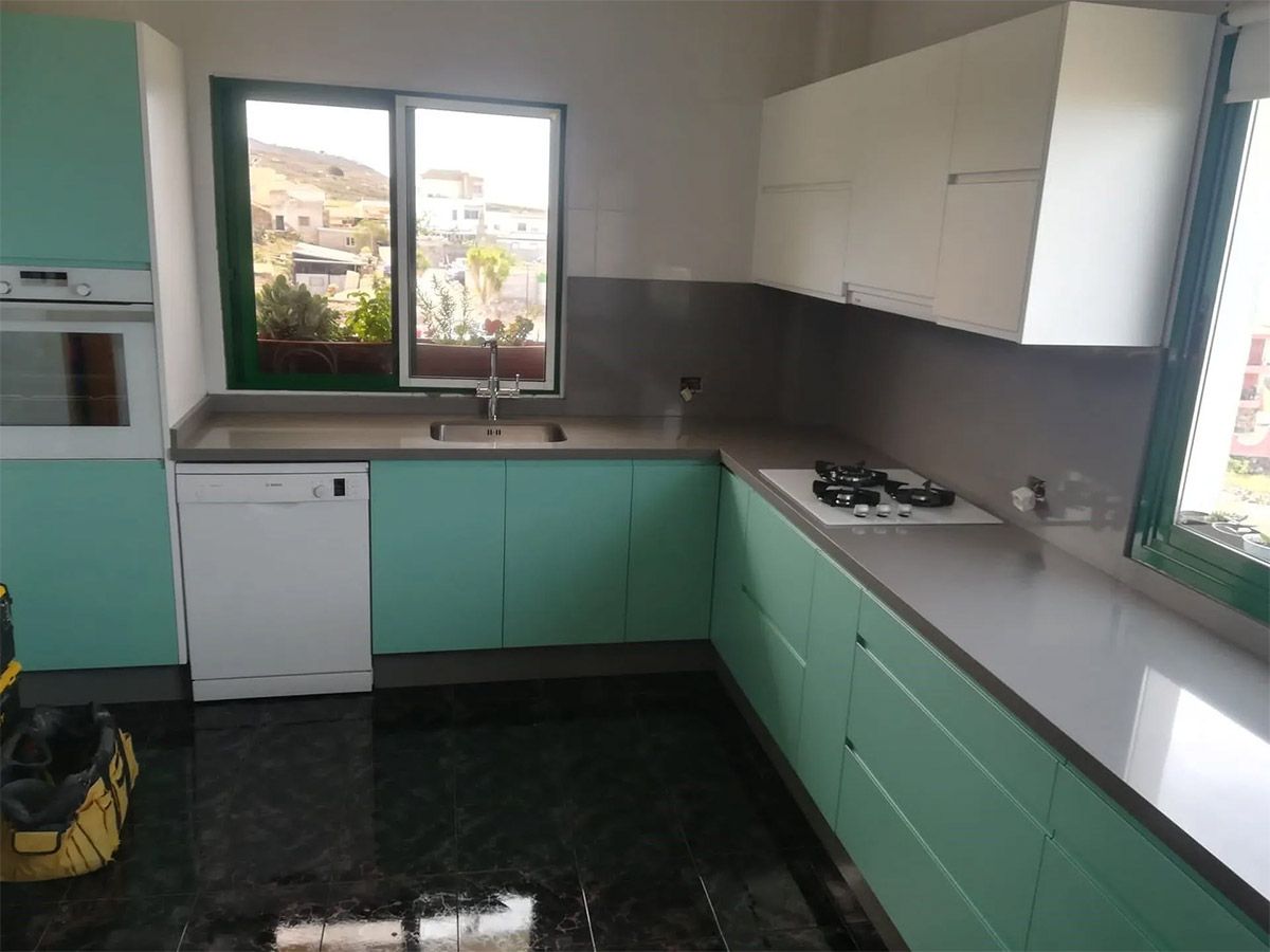 Cocina moderna vacía con armarios verde menta, encimera gris y luz solar que entra por dos ventanas.