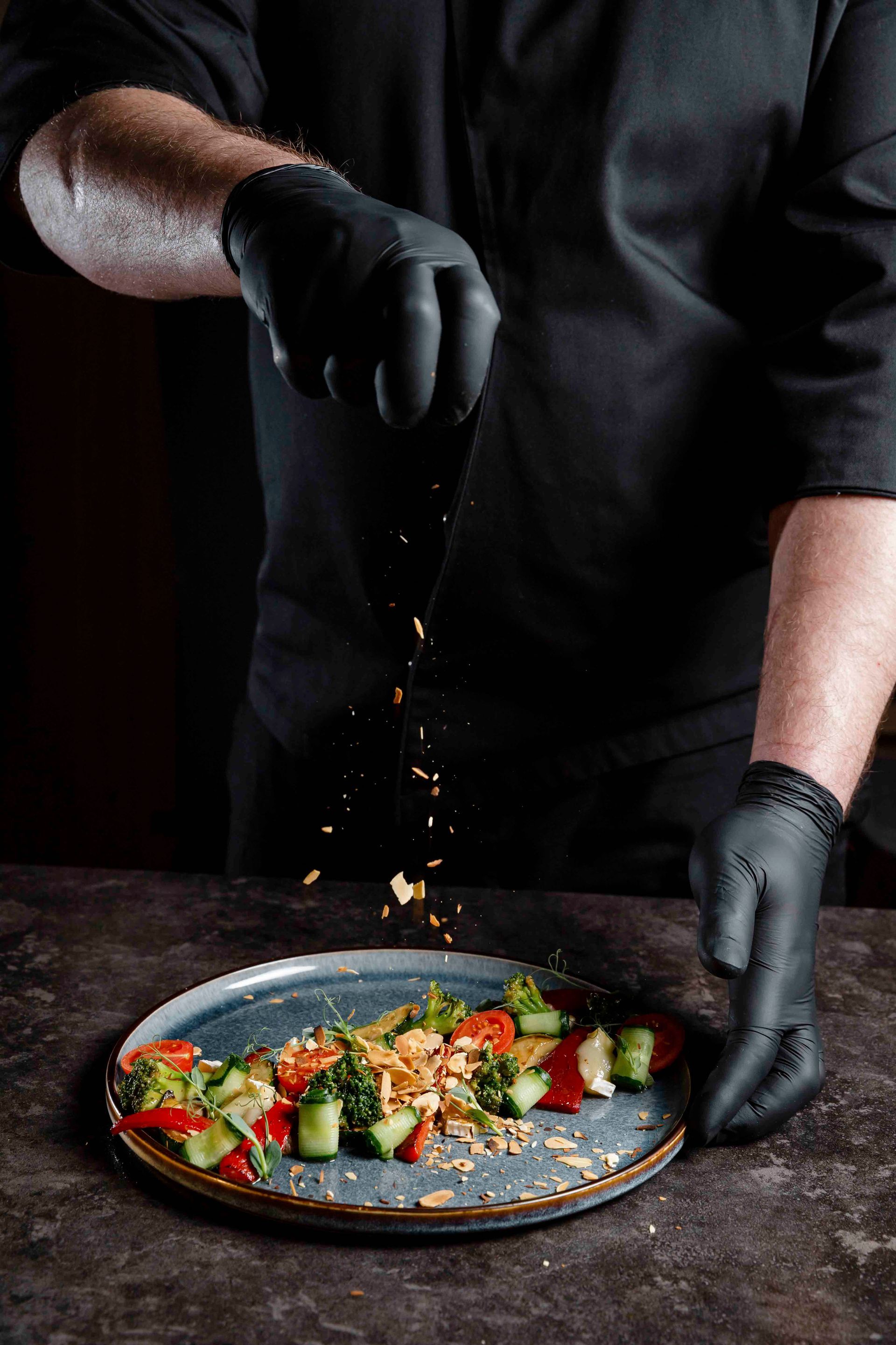 Koch in schwarzer Uniform streut Garnitur auf einen Salat auf einem grauen Teller.