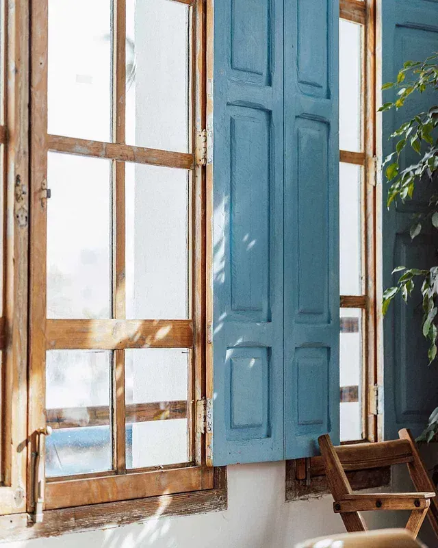 Una ventana de madera con contraventanas azules y una silla delante de ella.