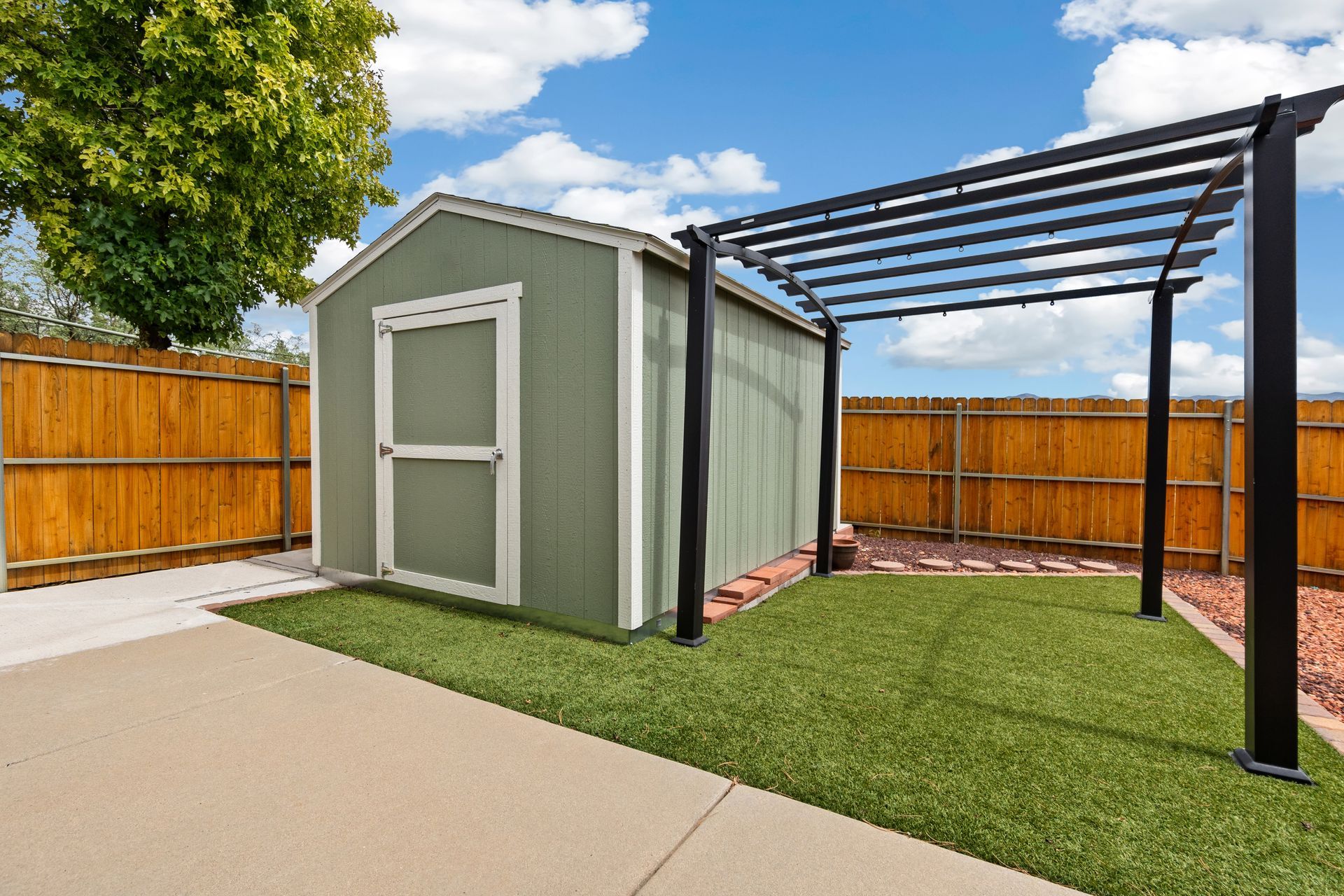 Abri de jardin avec pergola sur gazon synthétique, bordé d'une clôture en bois et d'une terrasse en béton.