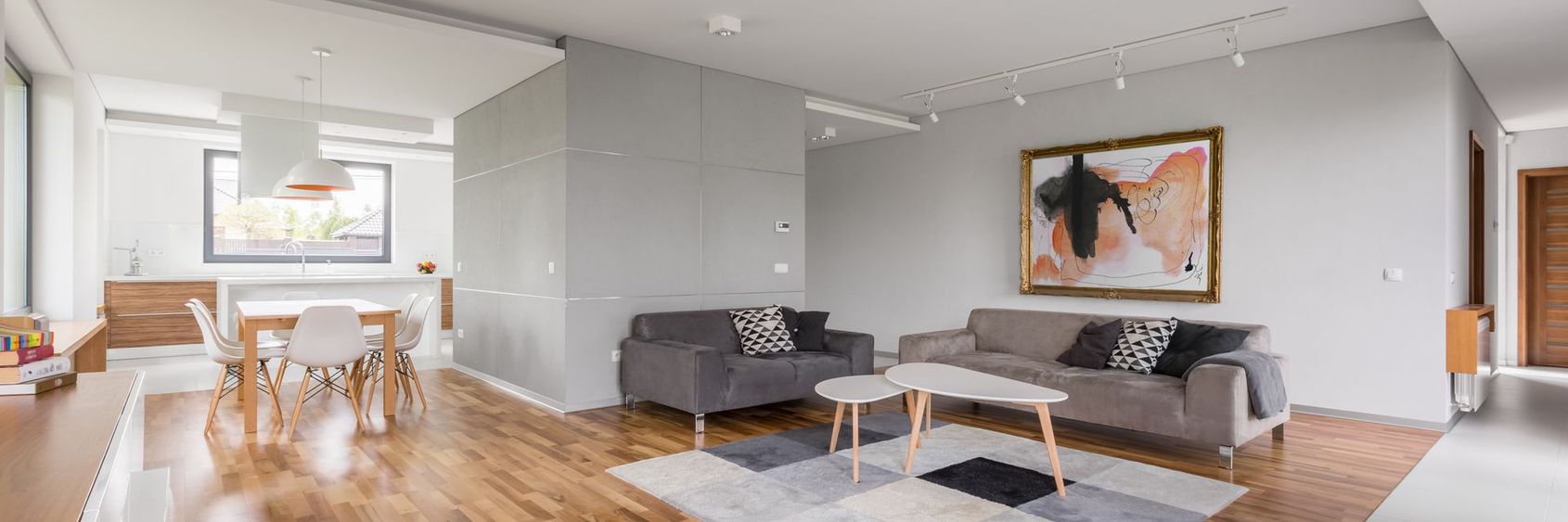 Salon moderne et lumineux avec canapés gris, murs blancs, parquet et une grande banquette près de la cuisine.