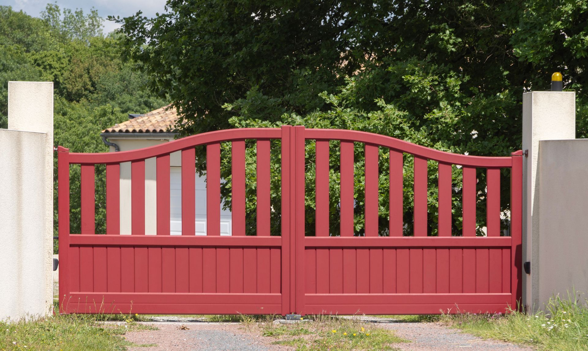 Un portail en fer forgé rouge à partie supérieure incurvée, entre des piliers de pierre, devant une maison et des arbres verts.