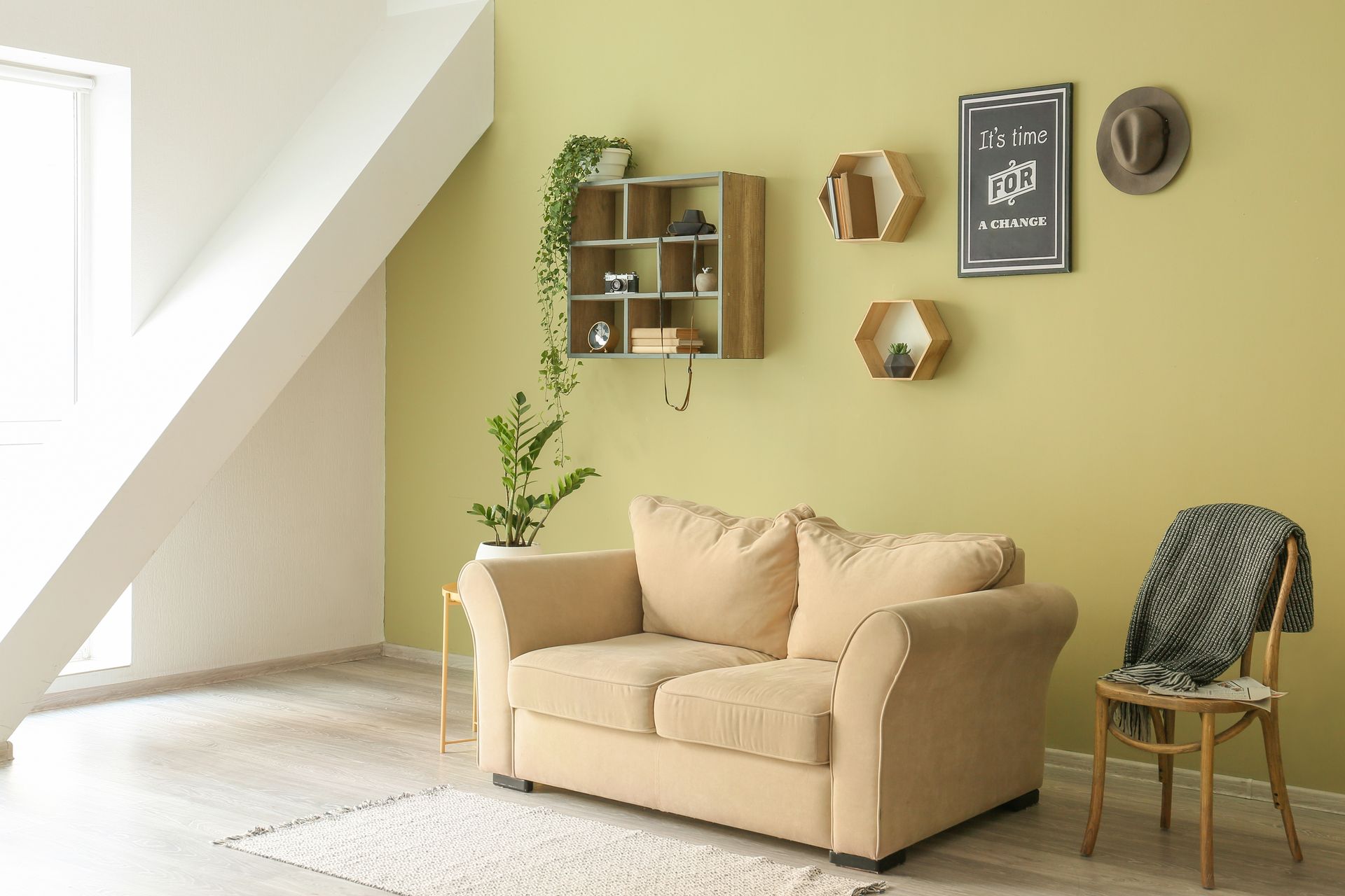 Salon beige cosy avec canapé, étagères murales, tableaux encadrés et fauteuil en bois près d'une fenêtre lumineuse.