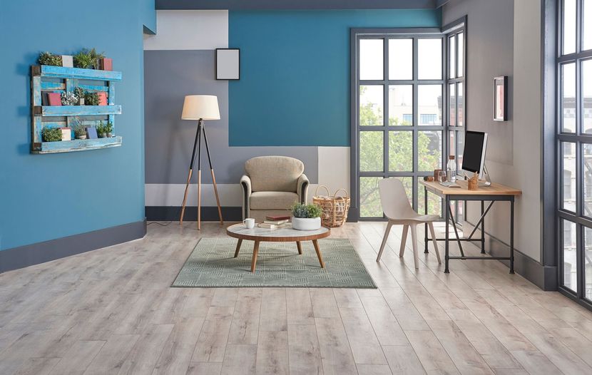 Salon moderne avec murs bleus, fauteuil gris, lampadaire, table basse et bureau près de la fenêtre.