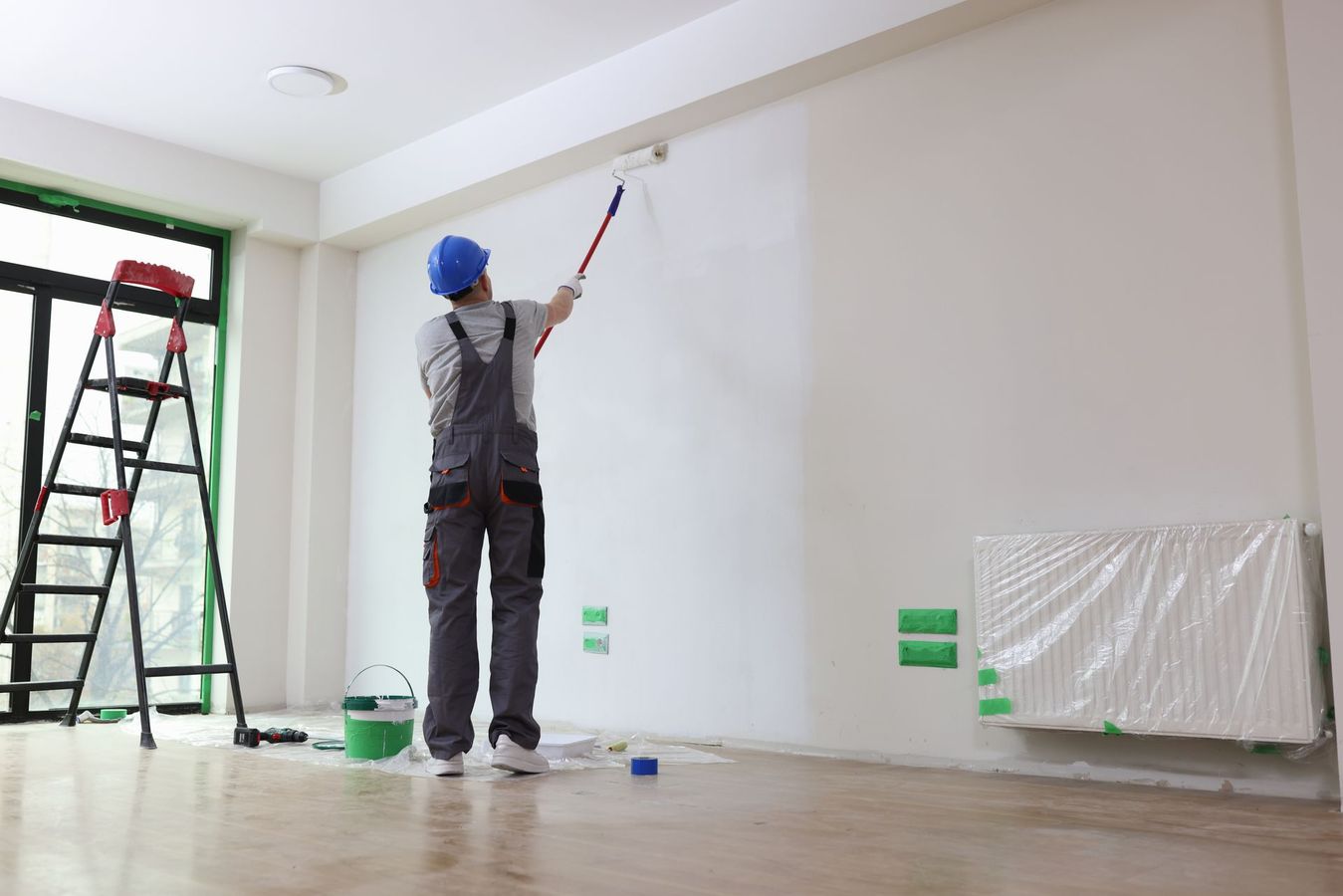Une personne peint un mur à l'intérieur avec un rouleau, à côté d'une échelle et de seaux de peinture.