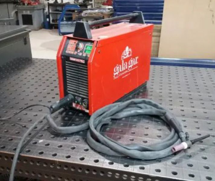 Maquina de soldar roja sobre mesa metálica, con cable y mango acoplados.