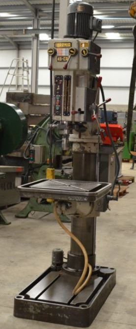 Un taladro de columna vertical en un taller. Es gris, con base rectangular y mesa metálica.