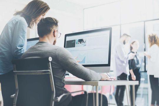 Dos personas mirando la pantalla de una computadora en una oficina; otras en el fondo.