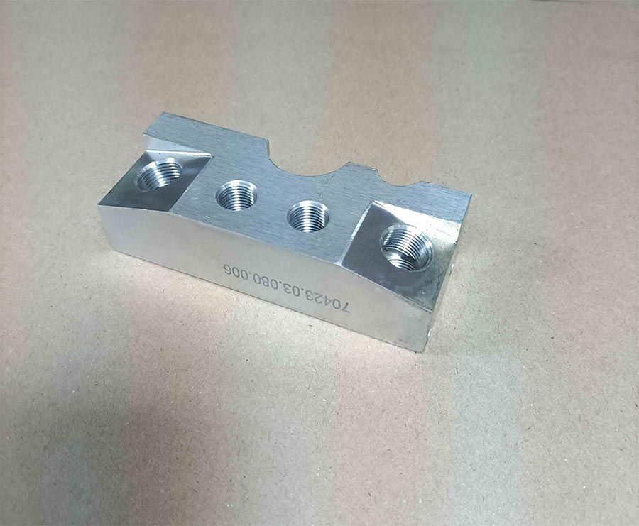 Bloque de metal plateado con tres agujeros y una ranura, sobre superficie clara.