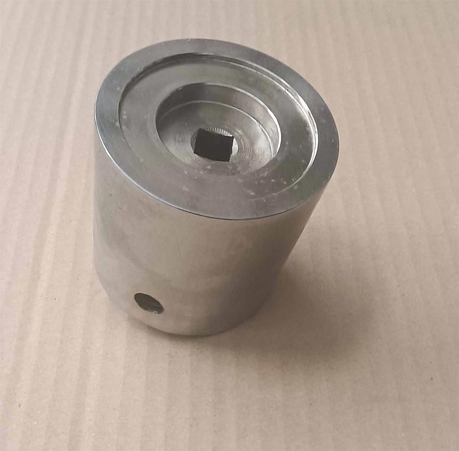 Herramienta cilíndrica metálica con un agujero cuadrado, una parte superior empotrada y un agujero lateral, sobre una superficie de cartón.