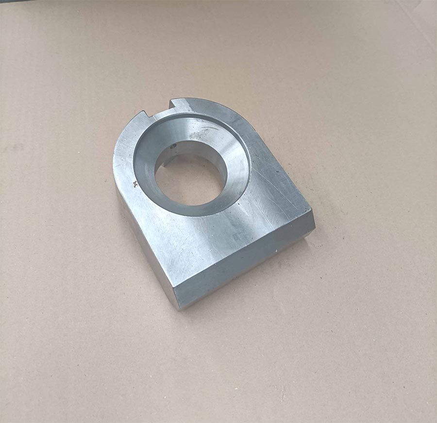 Pieza de metal plateado con orificio central y base rectangular, que reposa sobre una superficie de color beige.