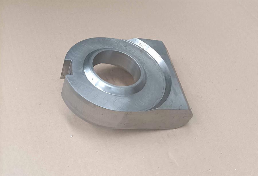 Pieza mecanizada de metal con apertura circular y bordes angulados sobre superficie ligera.