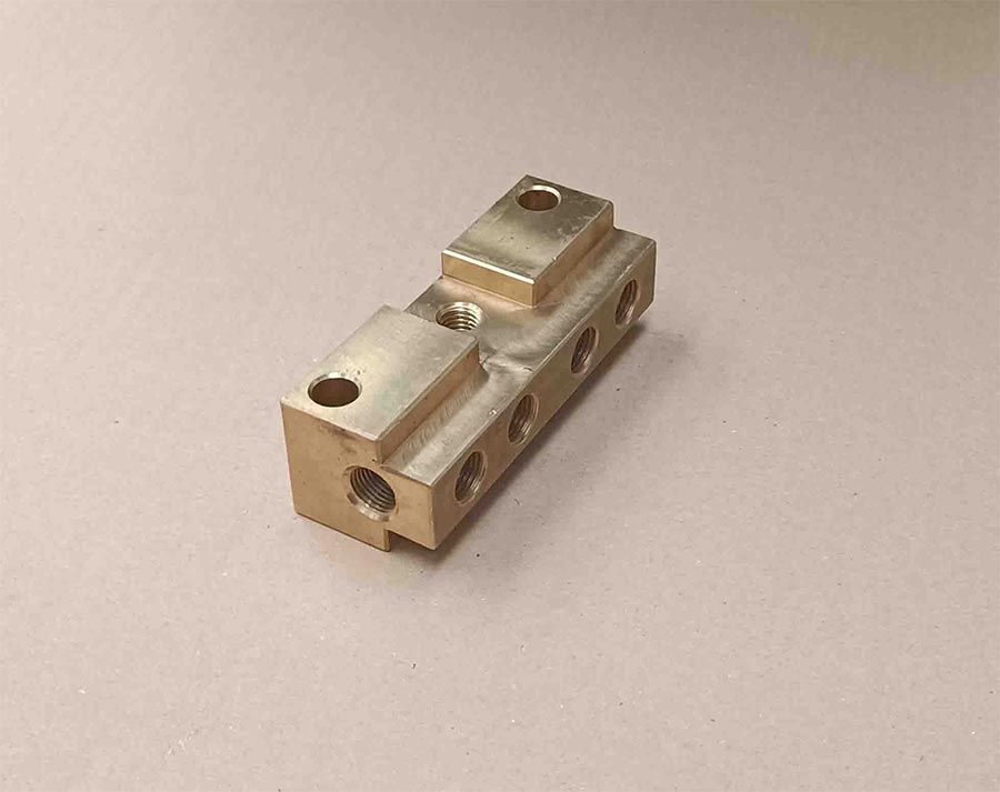 Bloque metálico de latón con agujeros y secciones elevadas, apoyado sobre una superficie neutra.