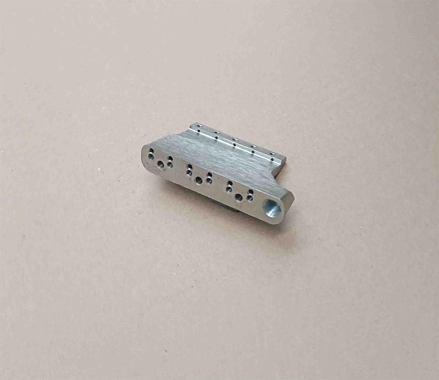 Componente metálico plateado con varios agujeros, sobre superficie neutra.