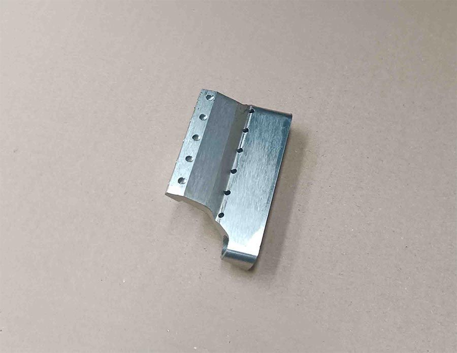 Soporte metálico con múltiples agujeros y cuchilla, sobre superficie ligera.